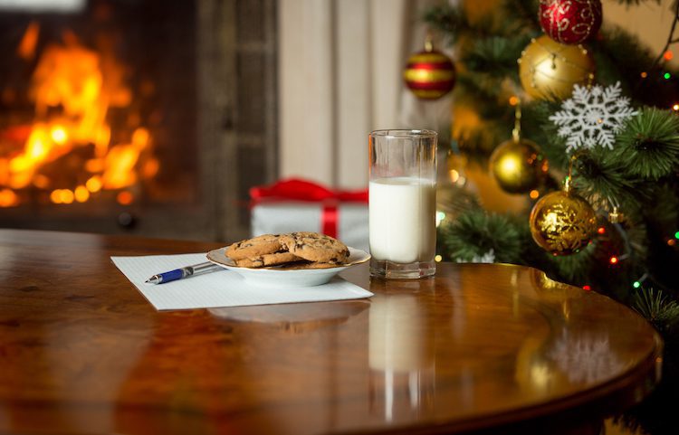 Closeup image of treats and letter to Santa on wooden table next Christmas tree and burning fireplace