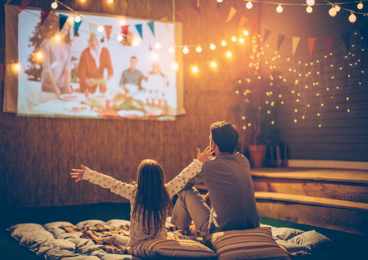 Father and daughter sitting at backyard and looking movie at home improved theatre. Backyard is decorated with string lighs.